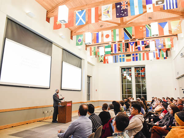 A man is speaking at a podium to a room full of people. Rows of flags from many different countries hang from the ceiling.